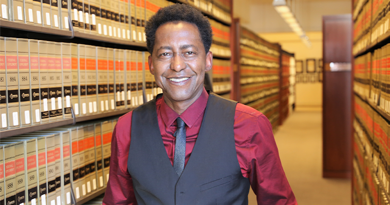 Image - Prof Kevin J Greene in red shirt and gray vest and gray tie standing in library aisle